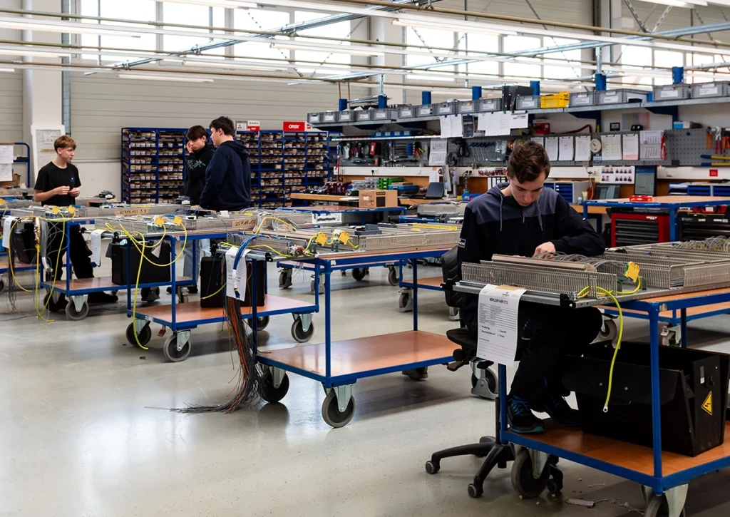 Mehrere Auszubildende arbeiten in einer modernen Werkhalle an Schaltschränken und elektrischen Komponenten.